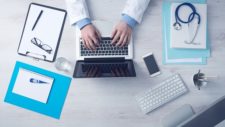 Someone working at a laptop, with a stethoscope, thermometer, and other supplies also scattered on the table.