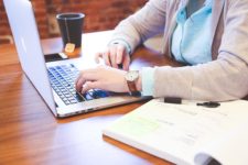 Someone sitting at a desk with a laptop, coffee, and open book, typing a case study.